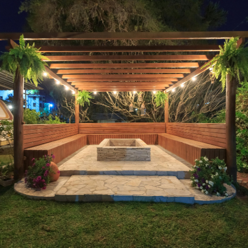 Wooden pergola with string lights and hanging ferns, surrounding a stone fire pit, with built-in wooden benches and landscaped flowers at night.